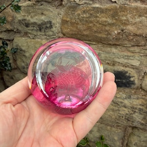 May include: A clear, round paperweight with a vibrant pink hue. The paperweight features an etched design of birds and floral patterns. The glass has a bubble-like texture, adding depth to the design. The paperweight is held in a hand.
