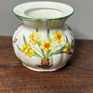 May include: A white ceramic vase with a green rim and a rounded body. The vase features a floral design of yellow daffodils, green leaves, and a brown base. The word "Narcissus" is written at the bottom.