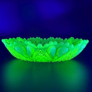 May include: A vibrant green glass bowl with a scalloped edge. The bowl features an intricate pattern of raised designs and a textured surface. It is lit with a bright green light against a dark blue backdrop.