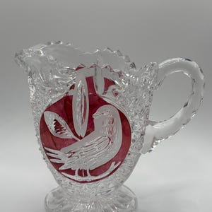 May include: Clear glass creamer with a decorative bird design etched on a red background. The creamer has a handle and a scalloped rim. The glass is cut with a diamond pattern.