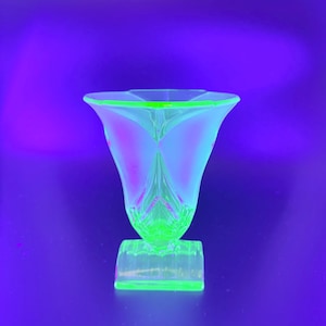 May include: A luminous green glass vase with a flared top and a square base. The vase has a distinctive shape with a design reminiscent of a flower. The glass has a subtle pink hue in the center, set against a purple backdrop.