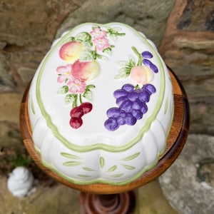 May include: A white ceramic serving dish with a fruit design, including peaches, grapes, and cherries. The dish has a scalloped edge with a green border and sits on a wooden pedestal.