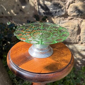 May include: A green glass cake stand with a circular design on top, supported by a silver metal base and a wooden pedestal. The stand is approximately 20 cm in diameter and is set against a natural background.