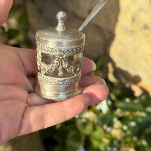 May include: A small silver-coloured condiment jar with a matching lid and a slender spoon. The cylindrical container features an ornate embossed floral pattern around the centre and a smooth base, with a clear glass insert visible between the metal bands.