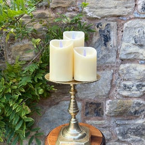 May include: Three ivory LED candles with flickering light effects on a gold metal candle holder. The holder has a decorative base and a round wooden stand. The background features a stone wall and green foliage.