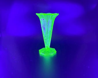 May include: A green glass vase with a fluted design. The vase has a flared top and a hexagonal base. The glass glows brightly against a dark blue background. The vase is likely vintage or antique.