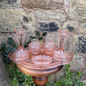 May include: A vintage pink glass vanity set, including two candle holders, two lidded jars, and a small dish, arranged on a tray. The set is displayed on a wooden pedestal, with a stone wall background.