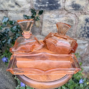 May include: A peach-coloured glass vanity set, comprising two perfume bottles and a rectangular tray. The set is displayed on a wooden stand against a stone wall. The bottles feature geometric designs.