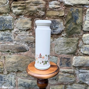 May include: A cream-coloured ceramic jar with a floral pattern, displayed on a small wooden stand. The jar is cylindrical with a lid and features a design of pink, yellow, and green flowers. The stand is dark brown and has a turned-wood design.
