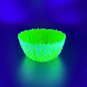 May include: A small, glowing green glass bowl with a scalloped rim and diamond and heart-shaped patterns. The bowl is illuminated against a dark blue background, creating a striking contrast. The bowl's design is symmetrical and detailed.