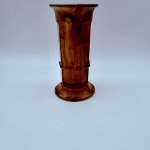 May include: A tall, cylindrical vase featuring a marbled brown and amber pattern throughout its surface. The vase has a flared rim at the top, a slightly tapered body with subtle vertical paneling, and a wide, circular base for stability.