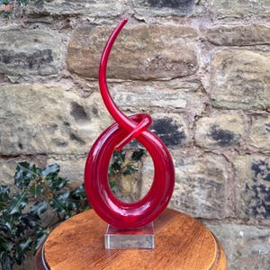 May include: A striking red glass sculpture featuring a looped design, presented on a clear acrylic base. The sculpture has a smooth, glossy finish and is displayed on a wooden pedestal against a stone wall. The artwork is a decorative piece.