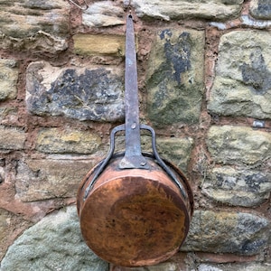 May include: An antique copper cooking pot with a long, riveted handle, suspended against a weathered stone wall. The pot features a rounded base and a dark, aged patina. The handle is made of iron and is attached to the pot with rivets, showing signs of age.