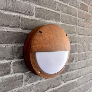 Puede incluir: Una lámpara de pared redonda para exteriores con un acabado metálico oxidado y desgastado. La lámpara tiene un difusor de luz blanco semicircular y está montada en una pared de ladrillo gris. La lámpara mide aproximadamente 20 cm de diámetro.
