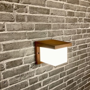 May include: A modern, square-shaped outdoor wall light fixture with a brown wooden frame and a white frosted glass panel. The light is mounted on a brick wall.