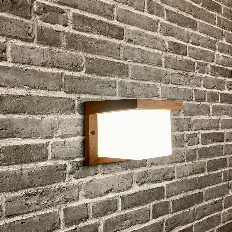 May include: A modern, rectangular wall light fixture with a wooden frame and a white frosted glass shade. The light is mounted on a brick wall.
