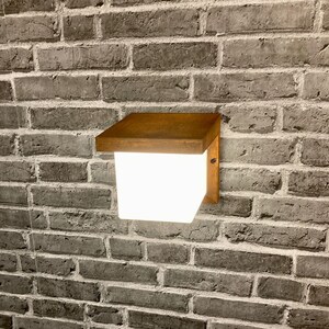 May include: A square, white light fixture with a brown wooden frame mounted on a brick wall. The light is on and illuminating the wall.