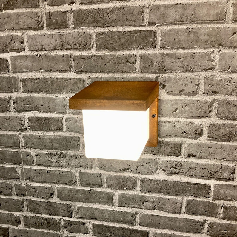 May include: A square, white light fixture with a brown wooden frame mounted on a brick wall. The light is on and illuminating the wall.