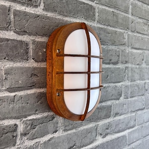 Puede incluir: Lámpara de pared exterior ovalada, marrón óxido, con interior blanco y barras metálicas. Montada en una pared de ladrillo gris, ofrece una estética rústica e industrial. Diseñada para uso exterior.