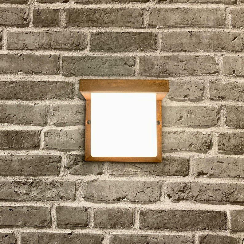 May include: A rectangular, wooden-framed light fixture with a white, frosted glass panel. The fixture is mounted on a brick wall.