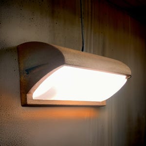 Puede incluir: Una lámpara de pared hecha de cerámica marrón con una pantalla de vidrio blanco esmerilado. La luz está encendida y proyecta un brillo cálido.