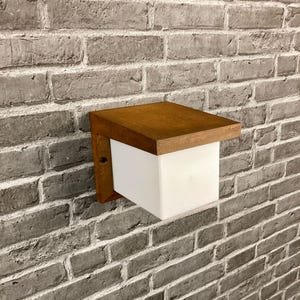 May include: A brown wooden outdoor wall light fixture with a white frosted glass shade. The light is mounted on a brick wall.
