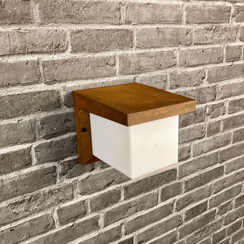 May include: A brown wooden outdoor wall light fixture with a white frosted glass shade. The light is mounted on a brick wall.