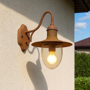 May include: An outdoor wall-mounted light fixture with a rustic, weathered appearance. The fixture features a curved arm, a round shade, and a clear, teardrop-shaped glass enclosure. The light is a warm yellow color, and the fixture is mounted on a beige wall.