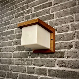 May include: A white square light fixture with a wooden frame mounted on a brick wall. The light fixture has a modern design and is perfect for outdoor lighting.