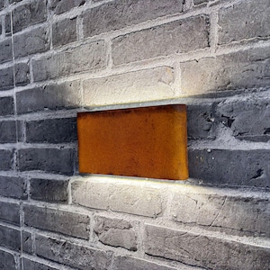 Könnte beinhalten: Eine rechteckige, rostorangefarbene Metallleuchte ist an einer grauen Ziegelmauer angebracht. Die Leuchte ist beleuchtet und wirft einen warmen Schein auf die umgebenden Ziegel. Die Ziegelmauer hat eine strukturierte Oberfläche.