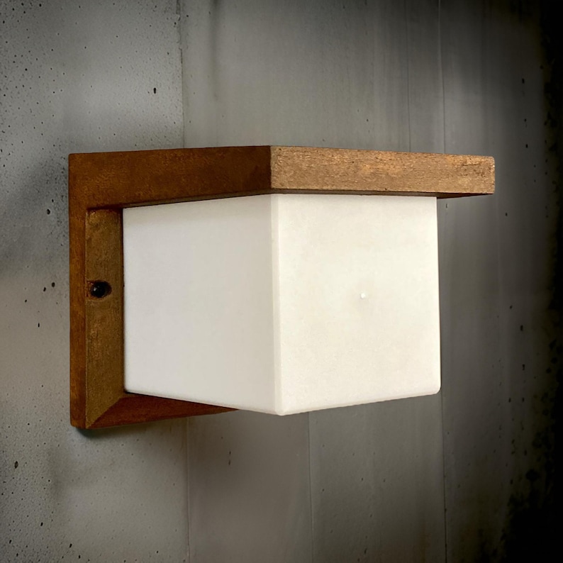 May include: A wall-mounted light fixture with a square, white frosted glass shade. The fixture is made of wood with a brown finish.