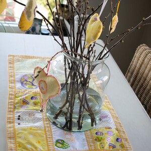 Op de afbeelding: Een glazen vaas gevuld met kale takken versierd met gele vilt hartjes en vogels. De vaas staat op een witte tafel met een geel en wit patchwork tafelkleed.