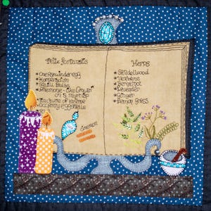 Op de afbeelding: Een gestikt boek met een blauwe achtergrond en witte stippen. Het boek is open om een lijst met kruiden en ingrediënten voor een drankje te onthullen. Het boek is versierd met kaarsen, een vijzel en stamper, en een kaneelstokje.