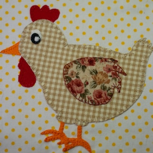 Op de afbeelding: Een bruine en witte geruite kip-applique met een bloemenpatroon op de rug. De kip heeft een rode kam en een rode keellap en oranje poten. De kip bevindt zich op een witte achtergrond met gele stippen.