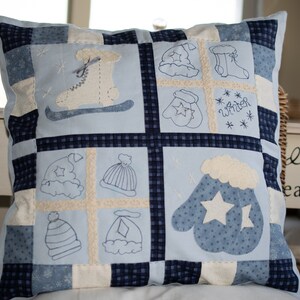 Op de afbeelding: Een blauw en wit patchwork kussen met geborduurde winterthema ontwerpen, waaronder wanten, mutsen, schaatsen en het woord "winter".