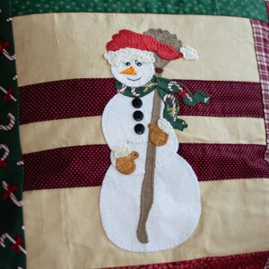 Op de afbeelding: Een witte sneeuwpop met een rode muts en een bruine stok in zijn hand. De sneeuwpop draagt een groen-witte sjaal en heeft zwarte knopen. De sneeuwpop staat op een beige achtergrond met rode en groene strepen.