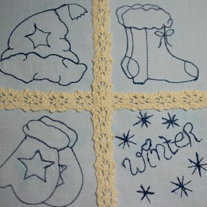 May include: A blue fabric square with four embroidered designs. The top left design is a blue and white hat with a star. The top right design is a blue boot with a white cuff. The bottom left design is a blue mitten with a star. The bottom right design is the word "winter" embroidered in blue with blue stars.