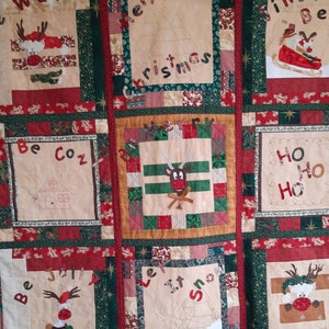 Può includere: Una trapunta patchwork beige e rossa a tema natalizio. La trapunta è composta da nove quadrati, ognuno con un design diverso. I design includono renne, alberi di Natale e le parole "Buon Natale", "Stare al caldo", "Essere allegri", "Lascia nevicare", "Ho Ho Ho" e "Caldi Jingle Bells".