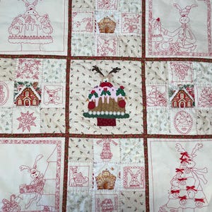 Op de afbeelding: Een wit quilt met rode borduursels die een kerstthema uitbeelden. Het quilt is gemaakt van negen vierkanten, elk met een ander ontwerp. De ontwerpen omvatten rendieren, peperkoekhuisjes, kerstboom en andere feestthema's.