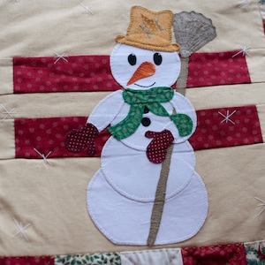 Op de afbeelding: Een witte sneeuwpop met een groene sjaal en rode wanten die een bruine bezem vasthoudt. De sneeuwpop staat op een beige achtergrond met rood-witte gepatenteerde stof.