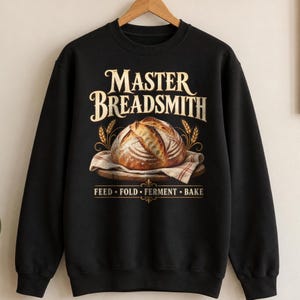 Puede incluir: Una sudadera negra de manga larga con un gráfico de una hogaza de pan redonda sobre una tabla de madera cubierta con un paño. El texto MASTER BREADSMITH aparece sobre el pan y FEED • FOLD • FERMENT • BAKE debajo en color dorado.