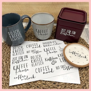 Könnte beinhalten: Ein blauer Keramikbecher mit dem Text DU.ICH.KAFFEE KLATSCH, eine weiße Keramiktasse mit KAFFEE Pause, eine dunkelrote quadratische Dose mit ZEIT FÜR EIN Käffchen, ein weißer Untersetzer mit coffee o'clock und ein Bogen schwarzer Aufkleber.