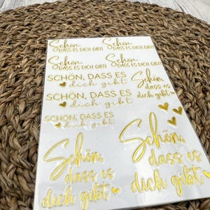 Könnte beinhalten: Eine Blatt mit goldenen Aufklebern mit dem Spruch "Schon, dass es dich gibt" (Deutsch für "Es ist schön, dass du da bist") mehrmals wiederholt. Die Aufkleber sind auf weißem Hintergrund und haben kleine Herzformen.