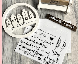 May include: White wooden sign with a circle cutout featuring a row of houses and the text "Herzlich Willkommen!" in black. The sign is surrounded by white paper with black text and images of houses, hearts, and leaves. The text includes phrases like "Zuhause ist, wo du bist!" and "Home sweet Home".