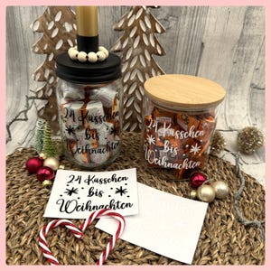 Könnte beinhalten: Zwei Gläser gefüllt mit verpackten Süßigkeiten, eines mit schwarzem Deckel und das andere mit Holzdeckel. Beide Gläser tragen den Text "24 Küsschen bis Weihnachten". Weihnachtsdekorationen umgeben die Gläser.