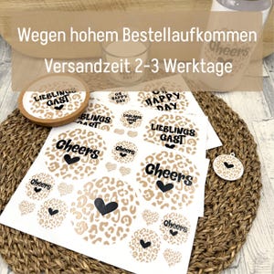 Könnte beinhalten: Ein Bogen mit runden Aufklebern in beigem und braunem Leopardenmuster. Die Aufkleber zeigen schwarzen Text wie "Cheers", "Lieblingsgast" und "Oh Happy Day", ergänzt durch kleine schwarze Herzsymbole. Ein Korkuntersetzer und ein kleiner Anhänger sind ebenfalls zu sehen.