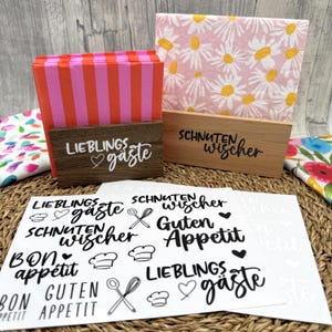 Könnte beinhalten: Zwei Serviettenhalter aus Holz mit Papierservietten. Ein Halter ist dunkelbraun mit dem weißen Schriftzug "LIEBLINGS gäste" und enthält rot-rosa gestreifte Servietten. Der andere ist helles Holz mit dem schwarzen Schriftzug "SCHNUTEN wischer" und Blumenservietten.