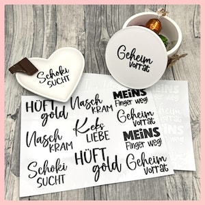 Könnte beinhalten: Ein weißes herzförmiges Gericht mit dem Text "Schoki Sucht" und einem Stück Schokolade. Ein weißer runder Behälter mit dem Text "Geheim Vorrat" und eine kleine Box mit einem Bonbon. Mehrere weiße Aufkleber mit verschiedenen deutschen Sätzen.