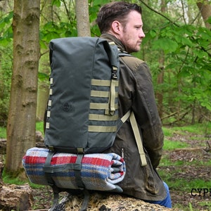 May include: A person wearing a green jacket and a large, dark green backpack with tan straps. The backpack has a red and blue plaid blanket strapped to the bottom. The person is sitting on a tree stump in a forest.