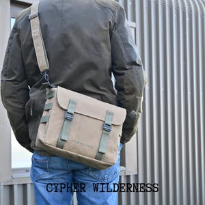 May include: A person wearing a green jacket and blue jeans is carrying a brown canvas messenger bag with green straps and buckles. The bag has the text "CYPHER WILDERNESS" printed on it.